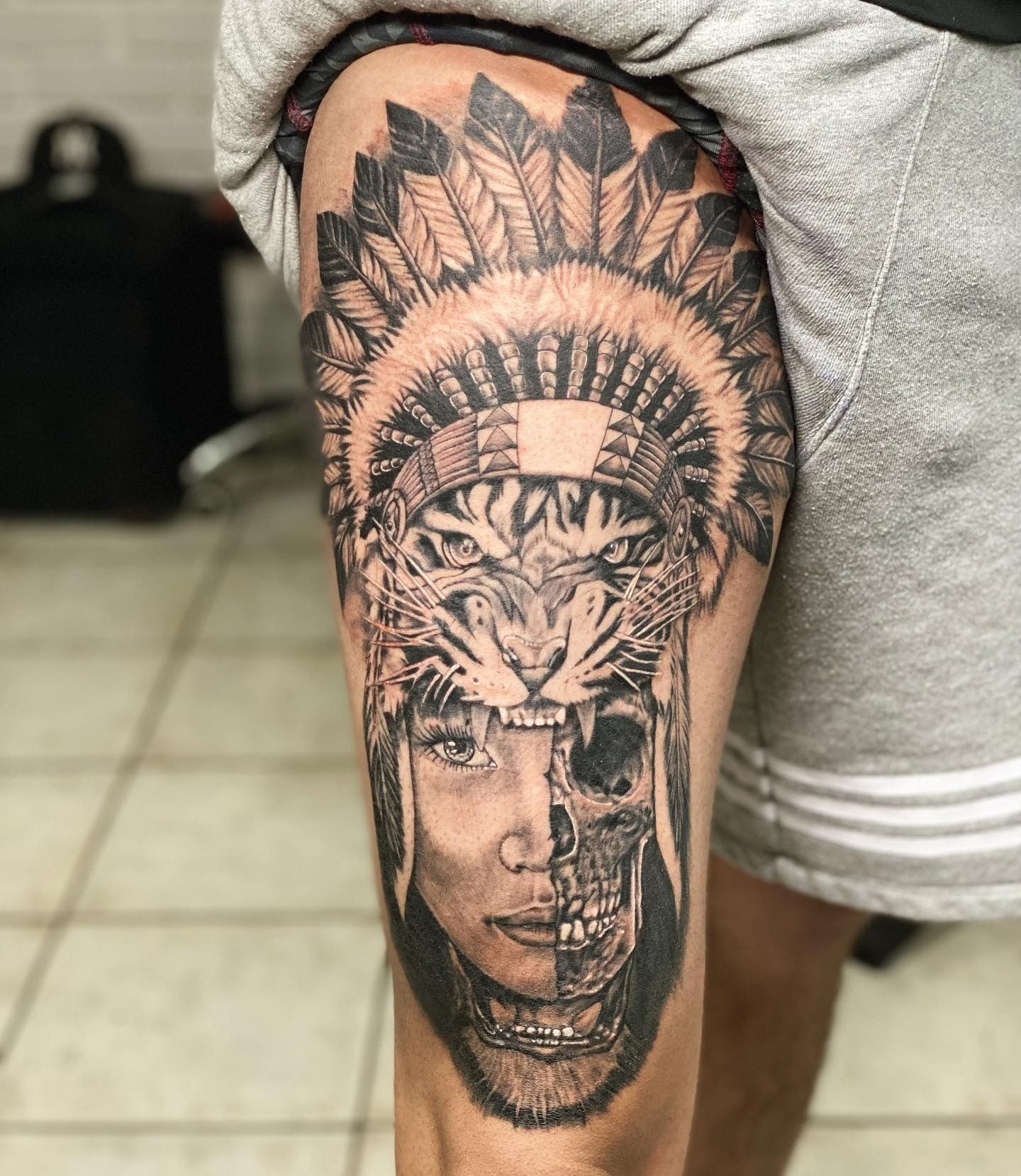 Tatuaje black & grey en el muslo: rostro de mujer con penacho de plumas, mitad tigre y mitad calavera