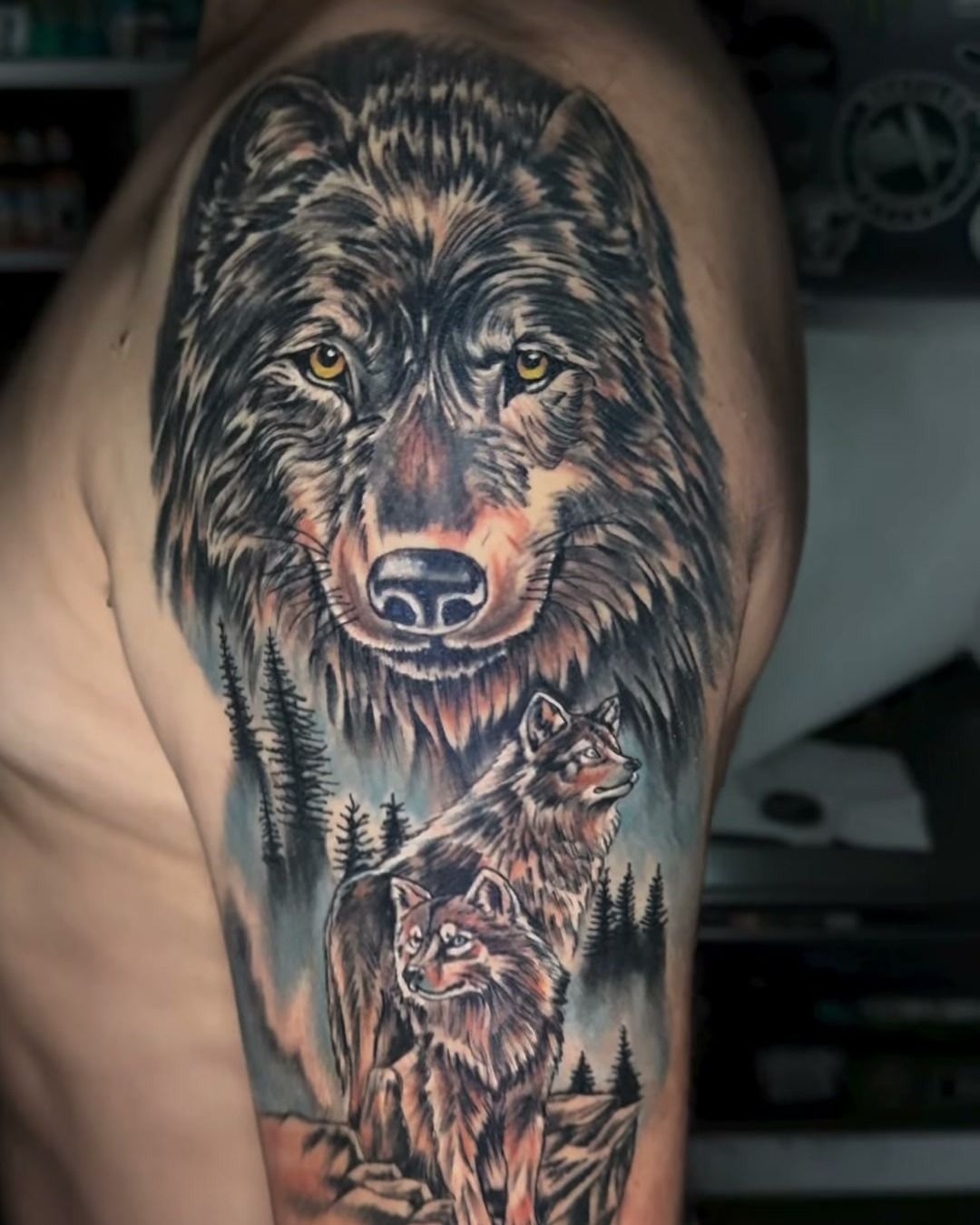 Tatuaje black & grey en el hombro: lobo de mirada ámbar sobre un bosque de pinos con una manada en la parte baja