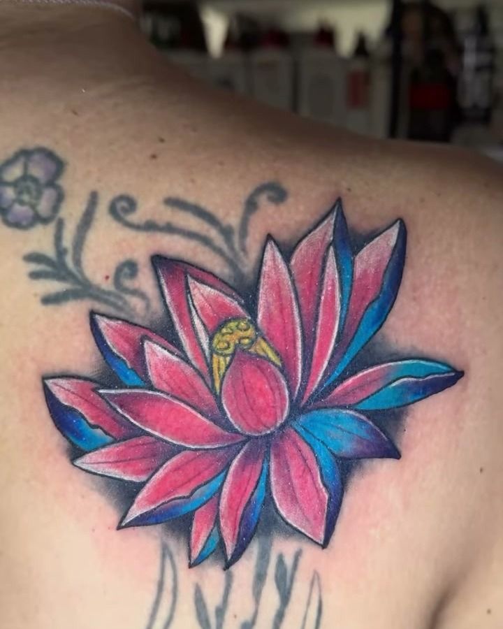 Restauración en el omóplato: flor de loto renovada en rosado y azul sobre un tatuaje floral antiguo